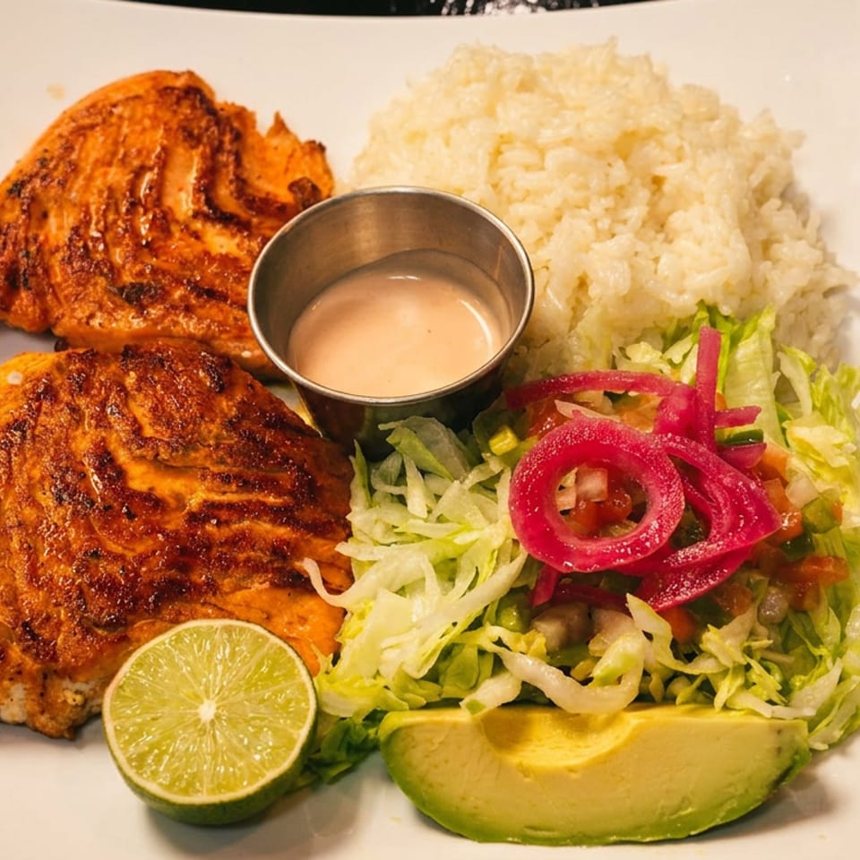 Grilled salmon with rice, salad, avocado, lime, and sauce on a white plate.
